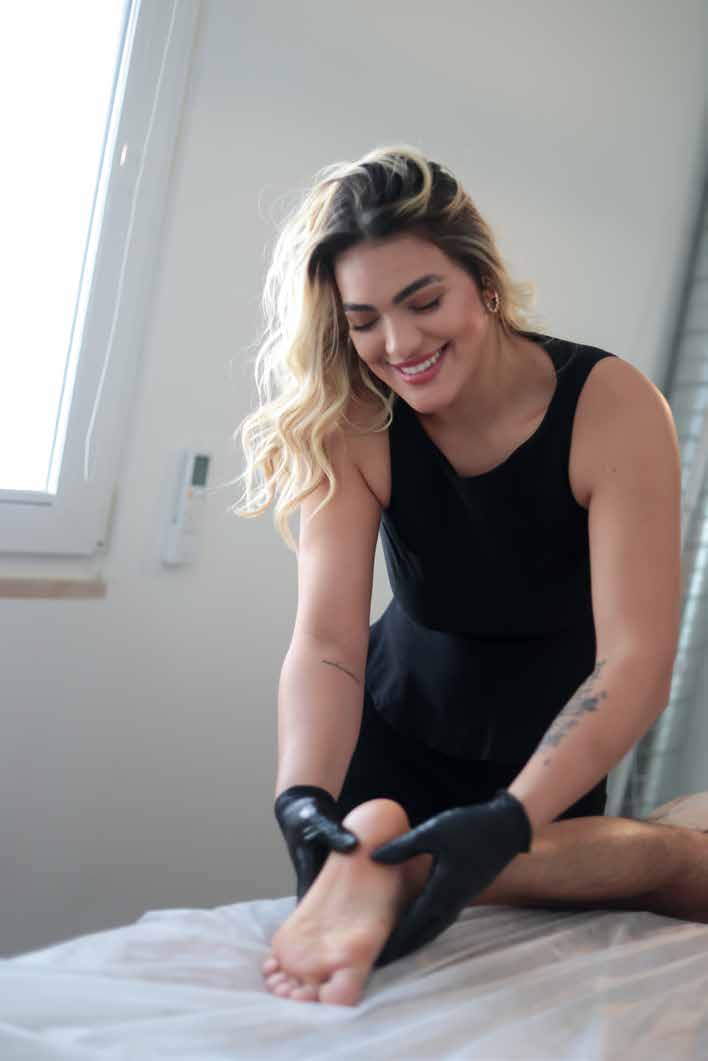Sabrina sorri enquanto trabalha no pé da cliente, ao lado da janela